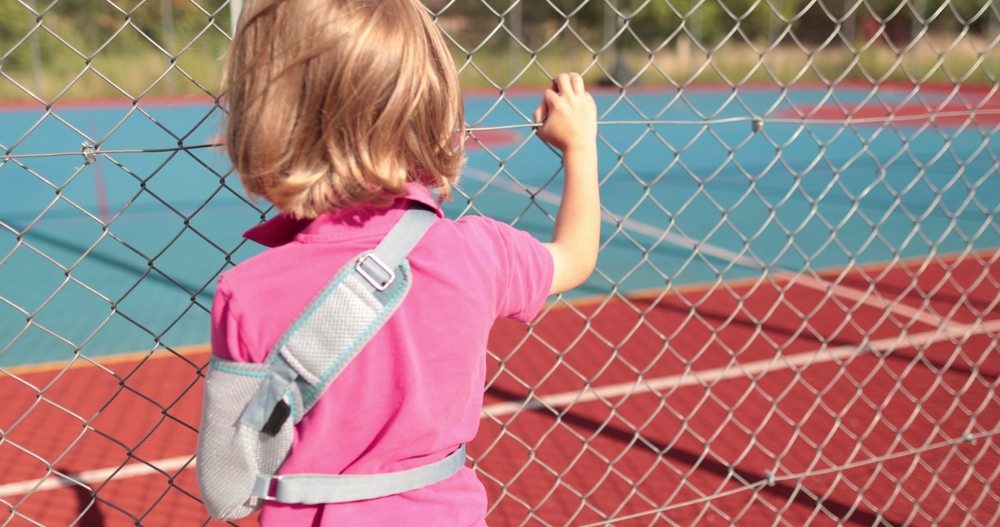 Child holding onto a chain-link fence looking toward a playground or sports court