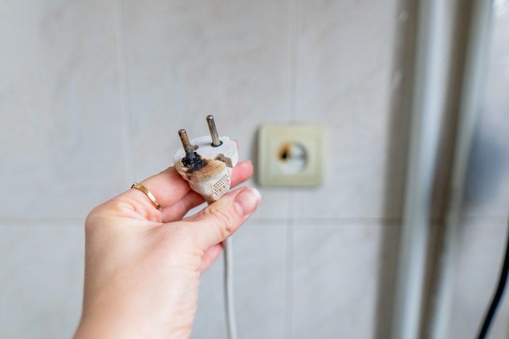 Hand holding a burnt and damaged electrical plug near a wall outlet