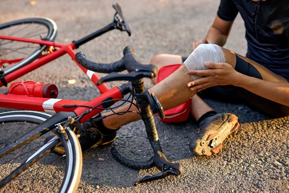 Cyclist sitting on the road holding a bandaged knee injury beside a fallen bicycle