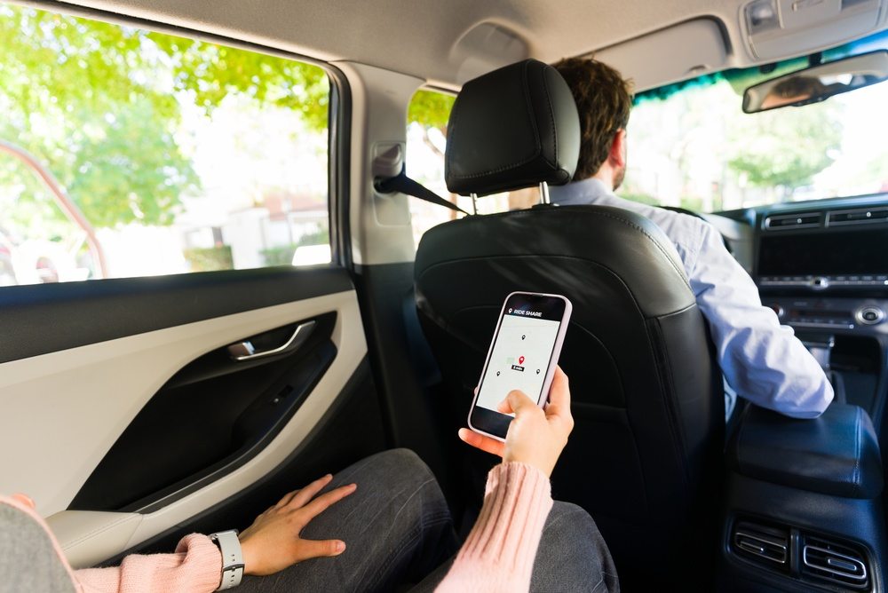 Rideshare passenger in the back seat holding a phone with a ride app map while the driver steers