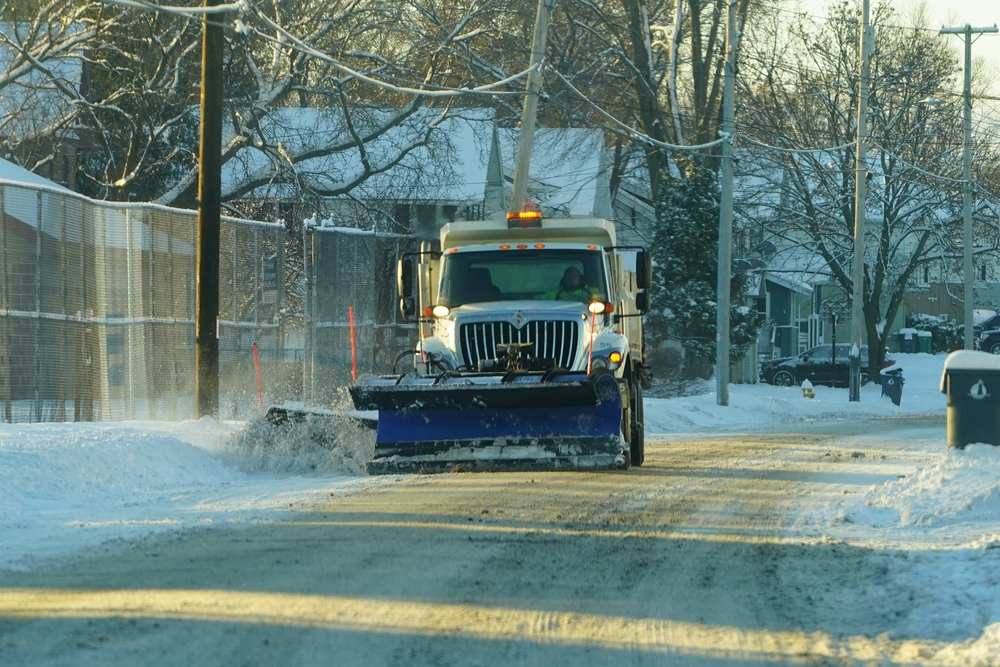 Municipal snow plow truck clearing a snow-covered residential street lined with houses and utility poles