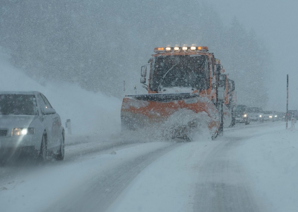 Orange snow plow throwing snow across a highway during a heavy snowstorm as a car passes in the next lane